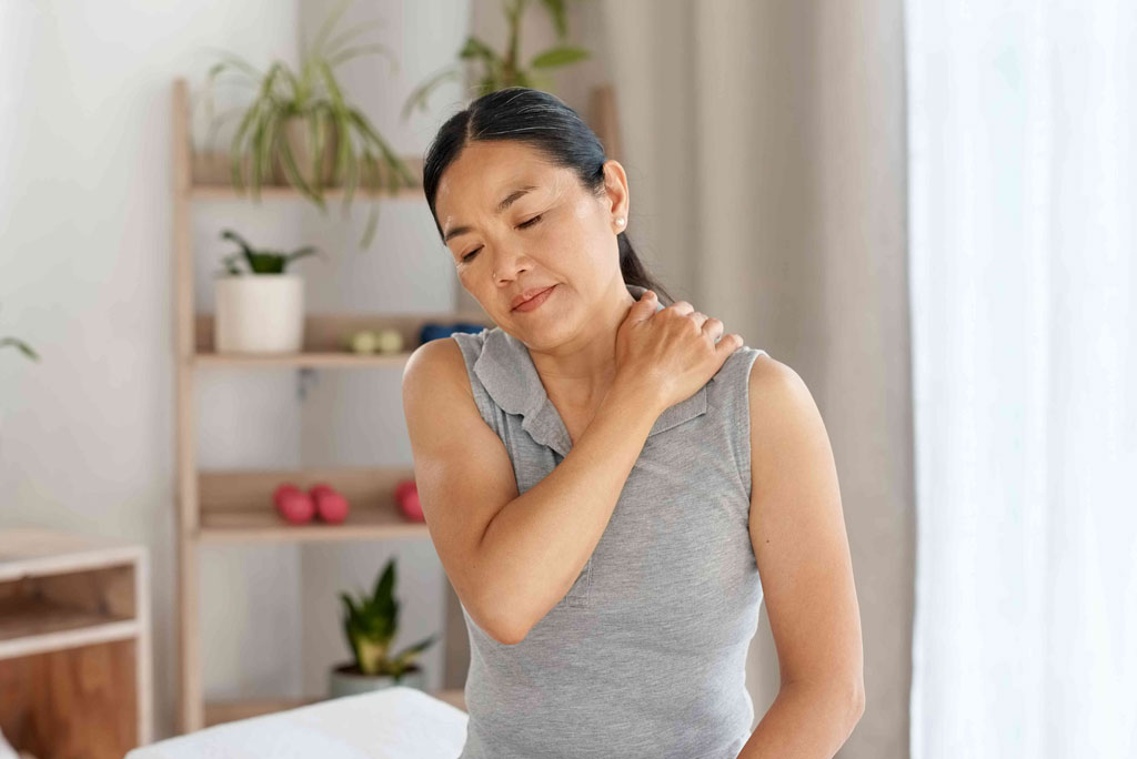 A woman massages her shoulder, experiencing pain; she is in a brightly lit room with plants and shelves.