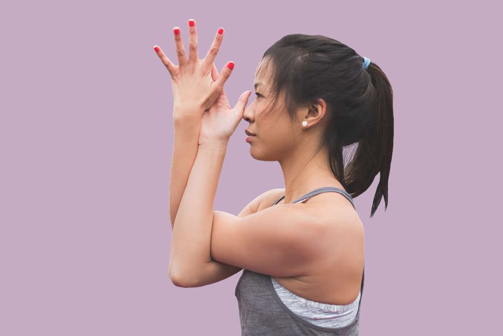 A woman performs a yoga pose, holding her hands together in front of her face, against a solid lavender background.