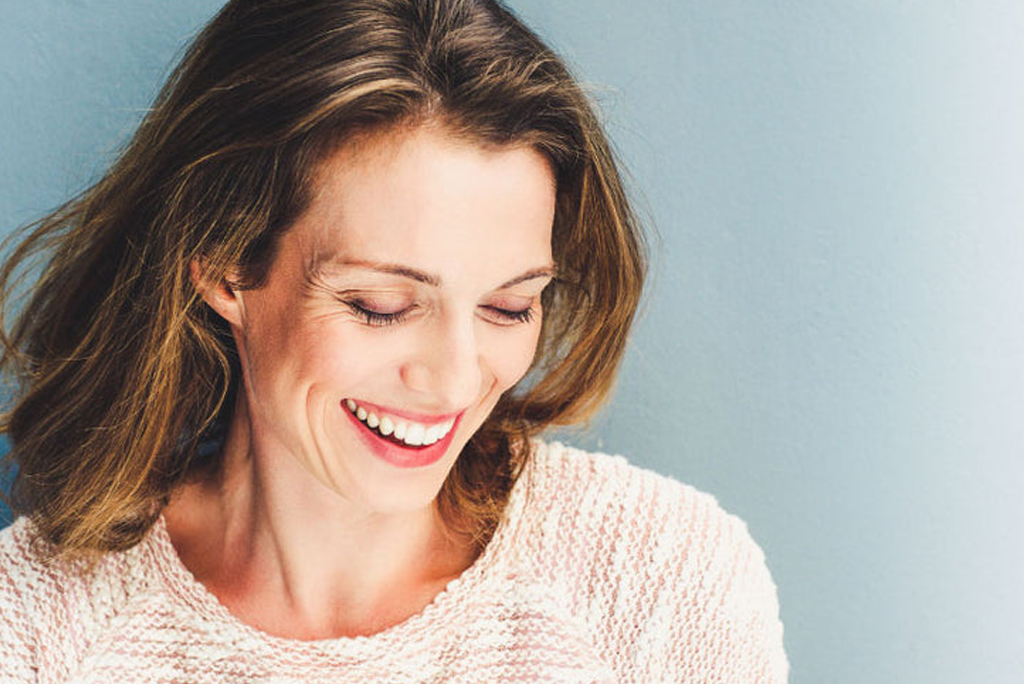 A woman with shoulder-length brown hair, smiles broadly while looking down, wearing a light-cream knitted sweater against a light-blue background.