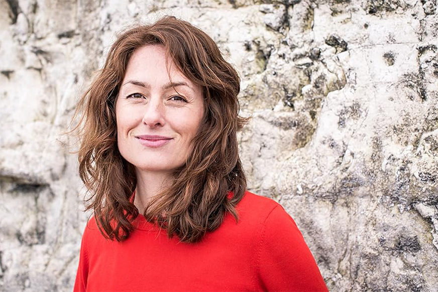 A woman with shoulder-length brown hair, wearing a red sweater, smiles subtly. She stands against a textured, light-grey stone wall.