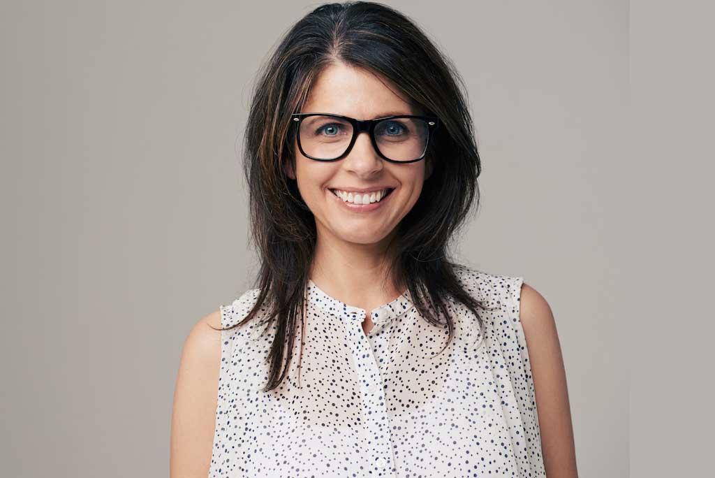 A woman with shoulder-length brown hair and glasses smiles directly at the viewer, wearing a white, patterned sleeveless top against a light gray background.