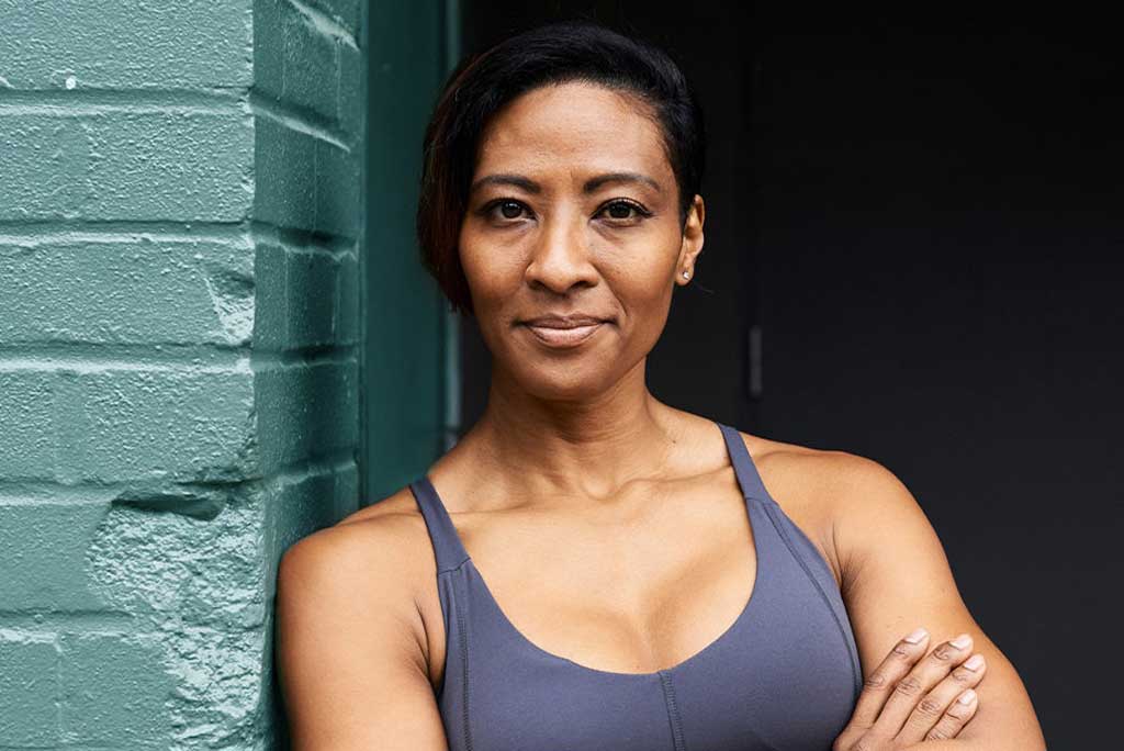 A woman with short dark hair, wearing a gray sports bra, stands with her arms crossed against a teal brick wall.