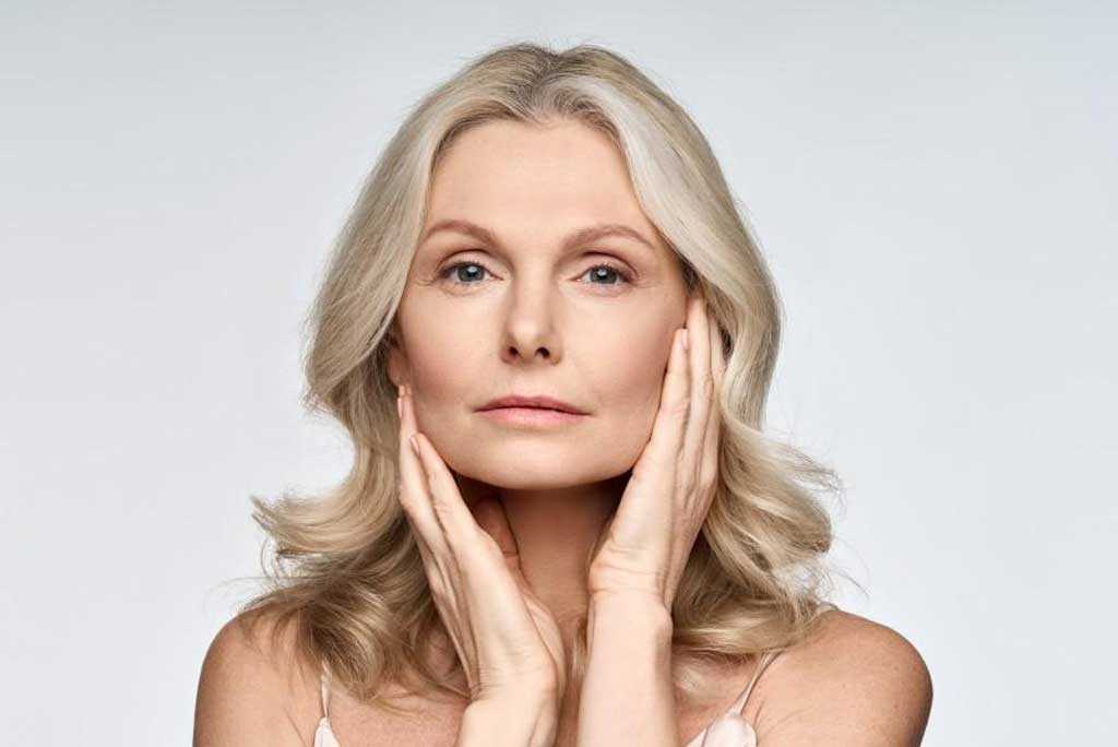 A woman with shoulder-length blonde hair gently holds her face with both hands. She is looking directly at the viewer against a plain light-grey background.